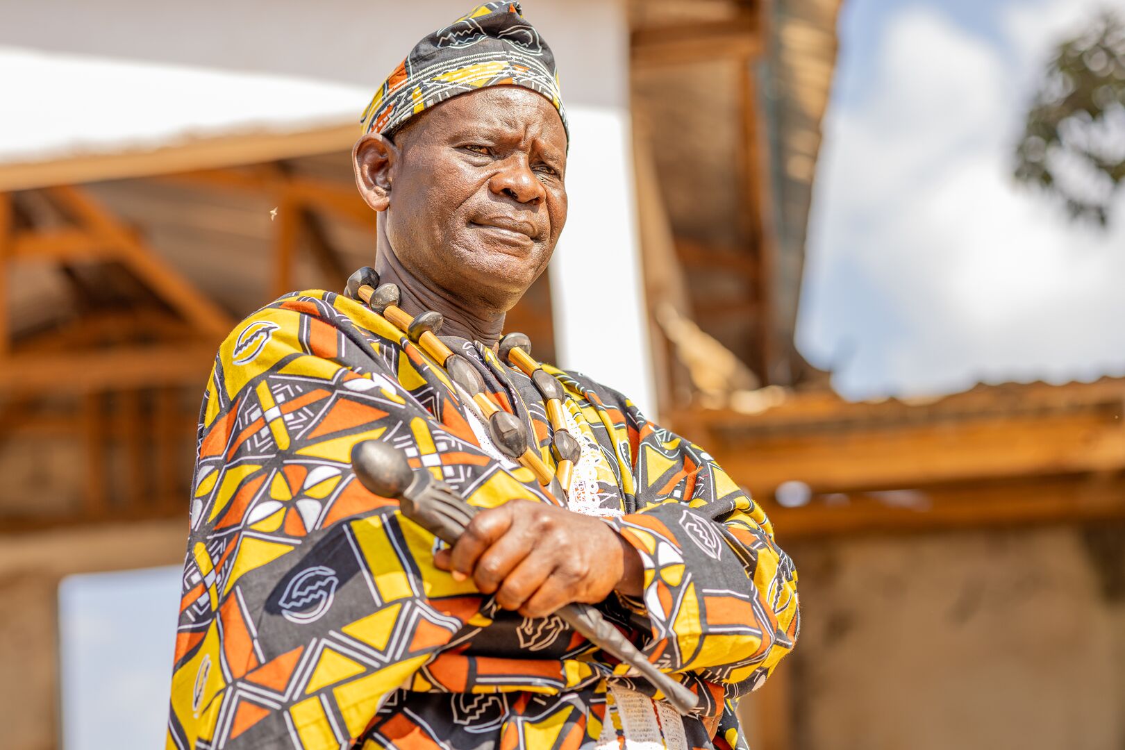 Chief Martin Azia Sodea stands facing the camera with his arms folded, in his village of Gado-Badzéré, Cameroon. He is wearing a brighlty patterned orange and yellow shirt and holding a metal staff.