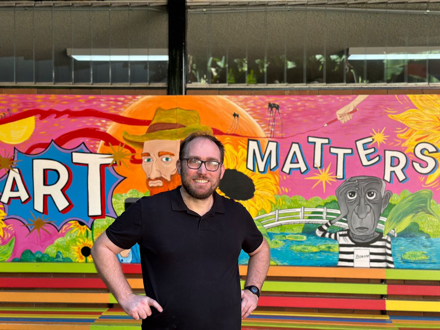 Isaac Owen, Director Of Treehouse Theatre Company, stands in front of a bright mural wall