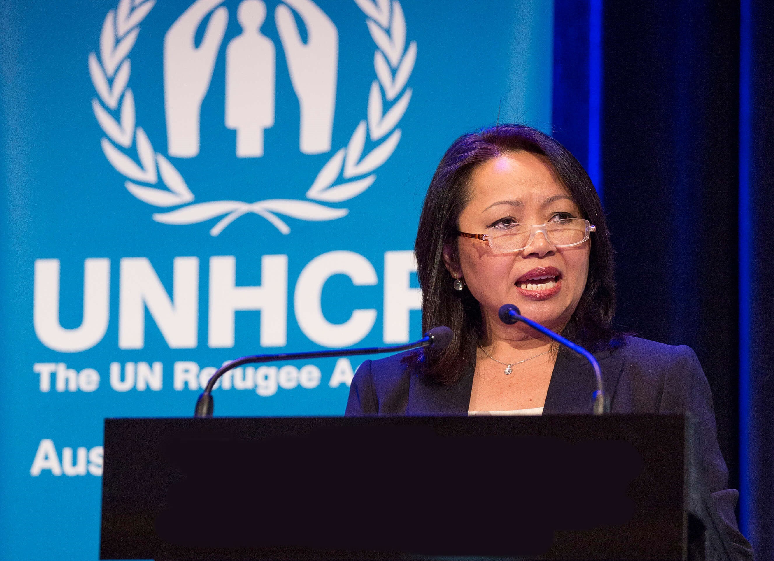 Special Representative Carina Hoang speaking behind a podium in front of Australia for UNHCR banner